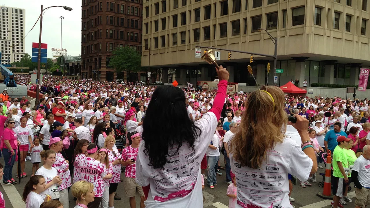 Two women on stage addressing a large crowd gathered for a charity walk or race event in a city, with one holding a bell and the other speaking into a microphone.