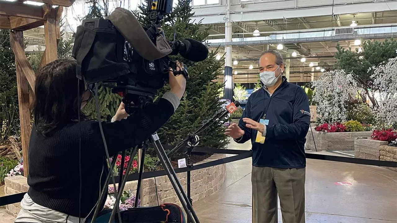 A man wearing a covid-19 mask being interviewed by a news crew in a greenhouse or botanical setting, surrounded by lush greenery and flowers.
