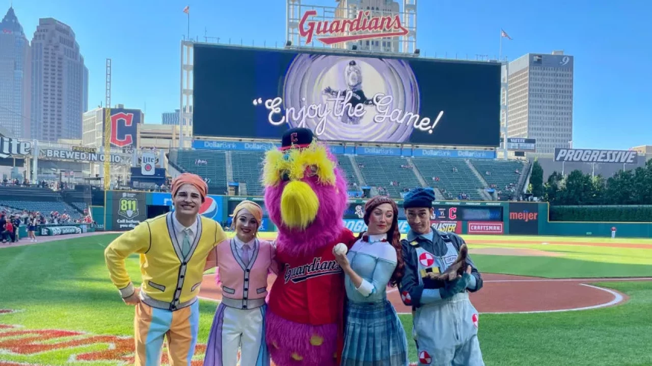 Performers posing with a brightly colored mascot on the baseball field at Guardians Stadium, with the scoreboard displaying the message 'Enjoy the Game!'