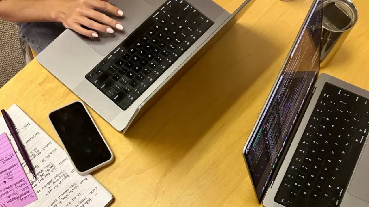 Close-up view of a workspace with two laptops, a notebook with handwritten notes, a phone, and a drink, capturing a productive setup for coding or studying.