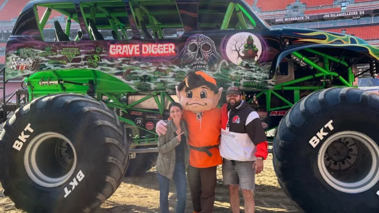 Two people posing with a mascot in front of the iconic Grave Digger monster truck at a stadium event.
