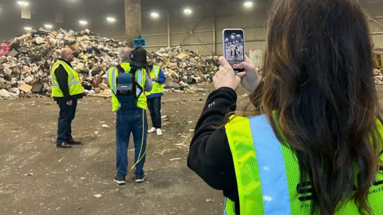 People in safety vests touring a recycling or waste management facility, with one person capturing the scene on their phone in front of large piles of sorted waste materials.