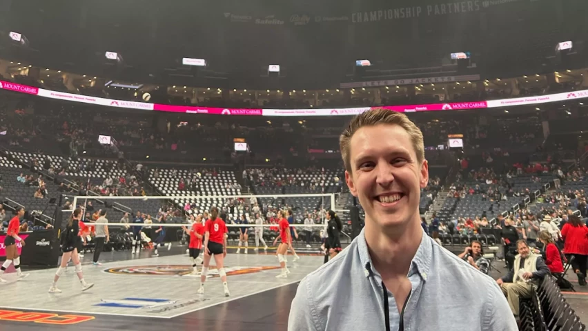 A smiling man standing courtside at a volleyball game, with players warming up on the court and a stadium audience in the background.